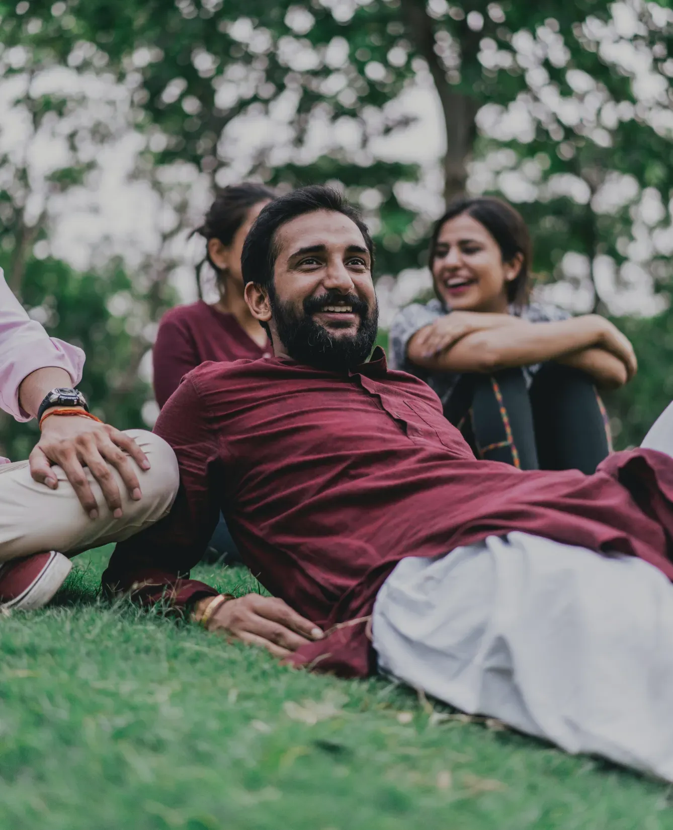 A man in a burgundy shirt and white pants leans back on a grassy field, smiling. Two people beside him also smile, creating a joyful atmosphere amidst lush greenery.