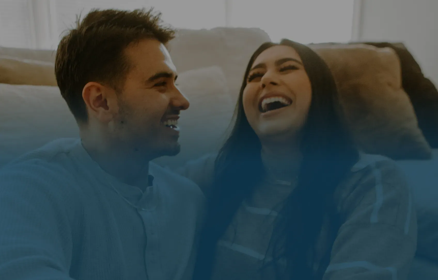 A couple sits on a couch, smiling warmly at each other. The woman is laughing joyfully. The scene conveys happiness and comfort in a cozy setting.