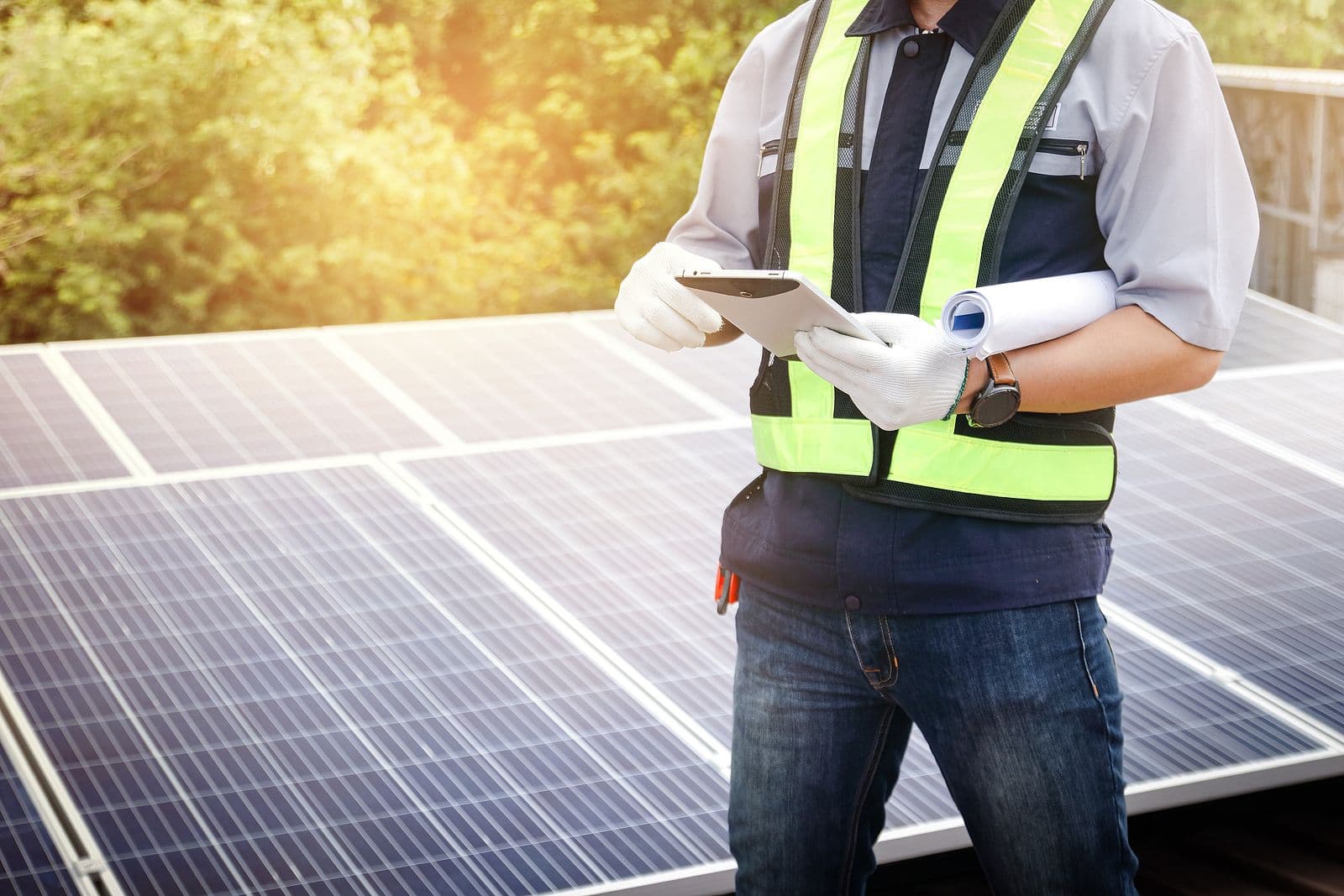 Inspector in safety vest and white gloves holding clipboard and rolled blueprint while inspecting rooftop solar panels