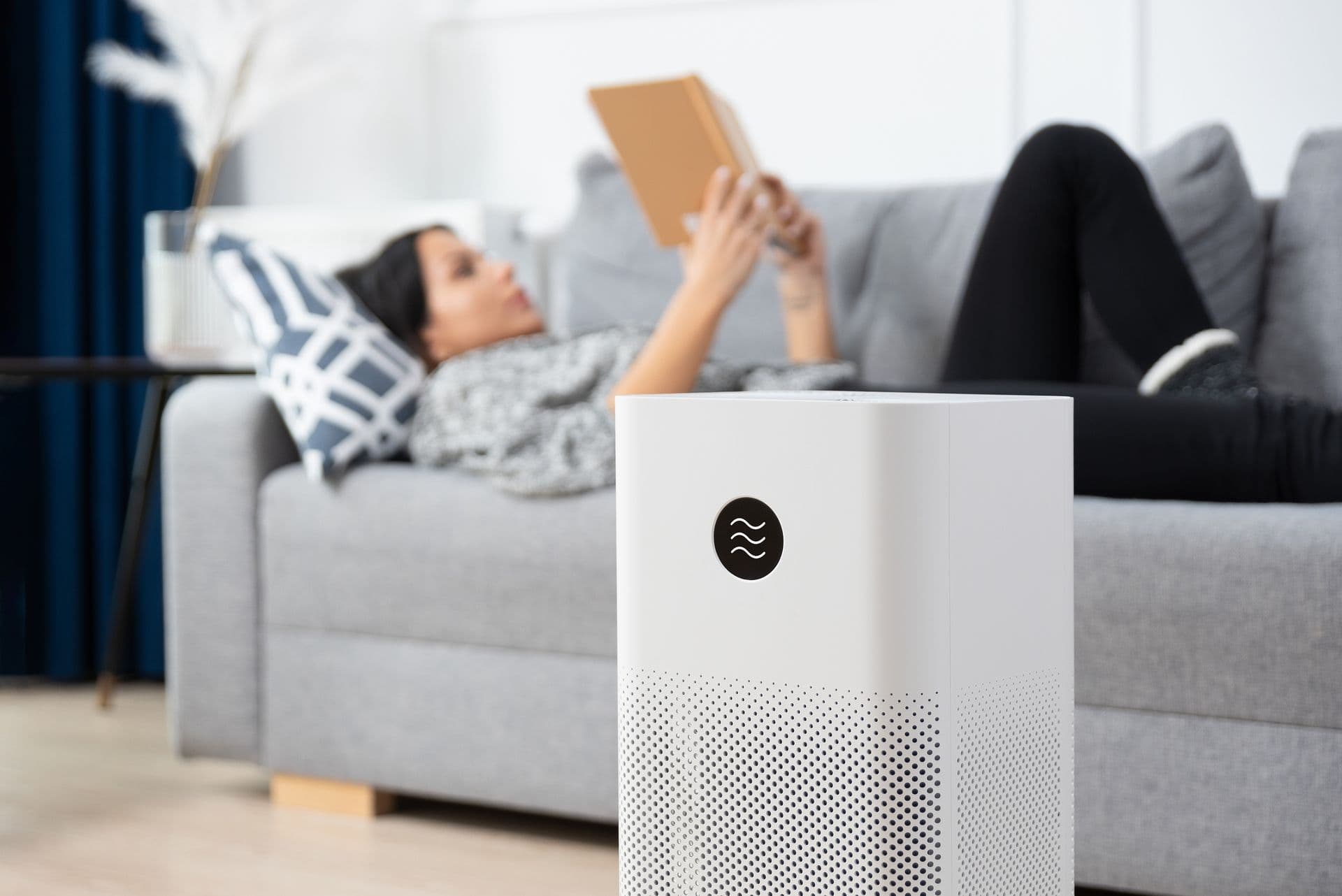 A white air purifier sits in the foreground of a modern living room. A person lounges on a gray sofa in the background, reading a book, creating a relaxed atmosphere.