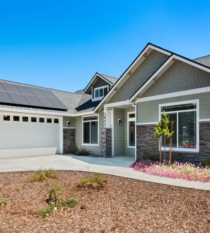 Single-story house with a gray facade and stone accents, solar panels on the roof, a two-car garage, and a small garden with flowering plants. Bright, sunny day.