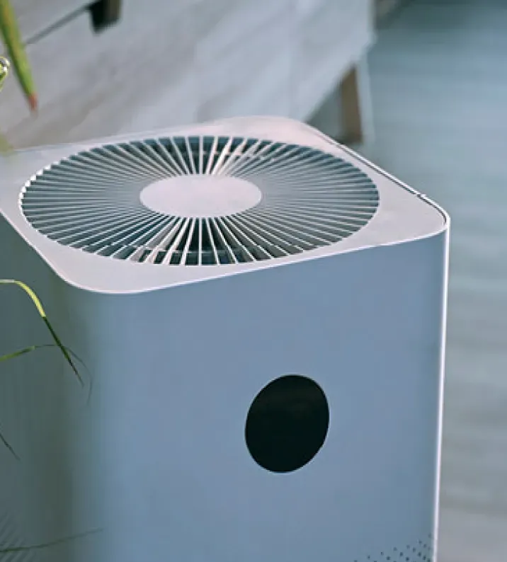 A sleek, modern air purifier with a circular vented top and a black sensor on the side, placed in a bright room with minimalistic decor.