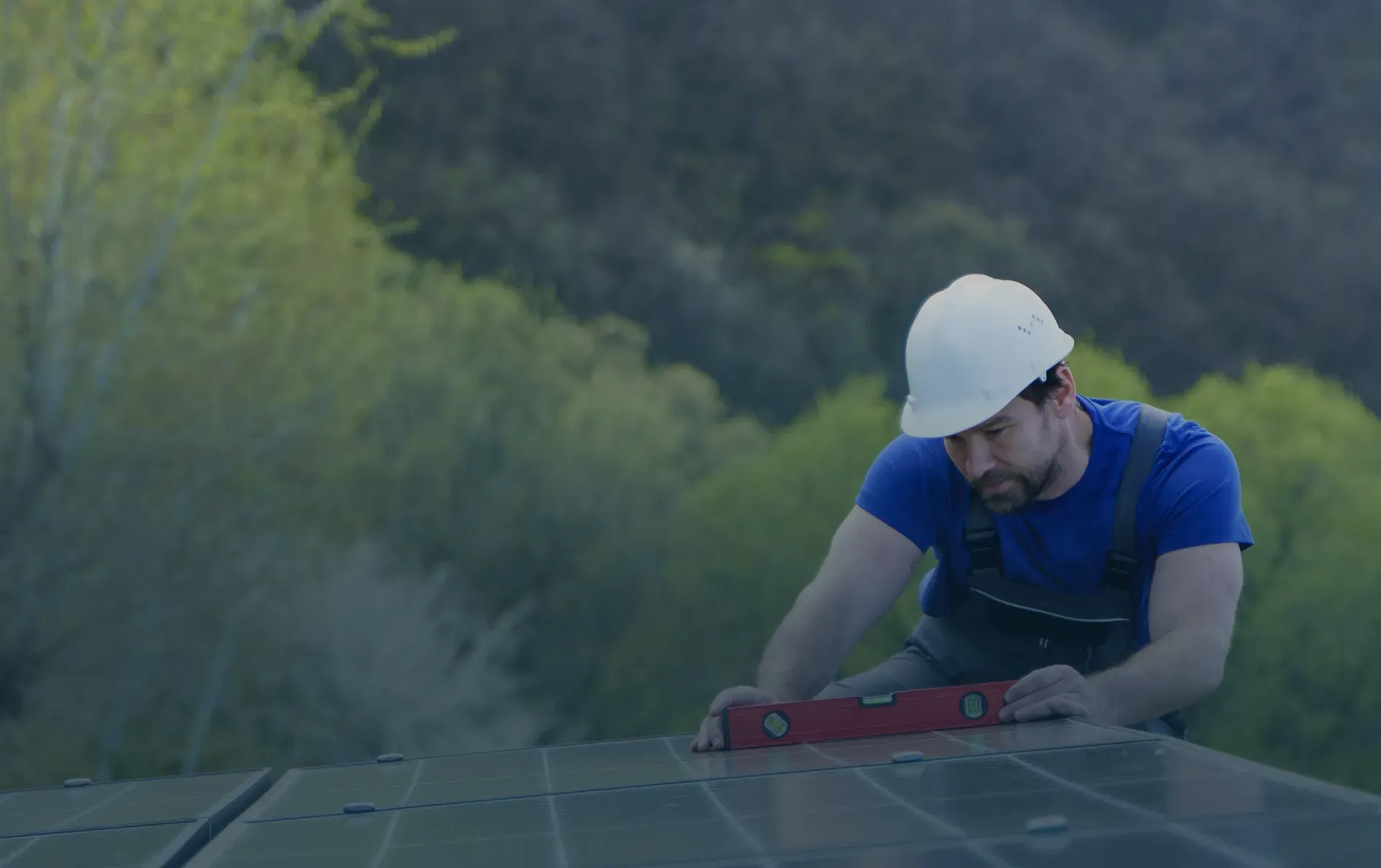 A worker in a blue shirt and white hard hat uses a level on solar panels, set against a backdrop of lush green trees. The scene conveys focus and dedication.