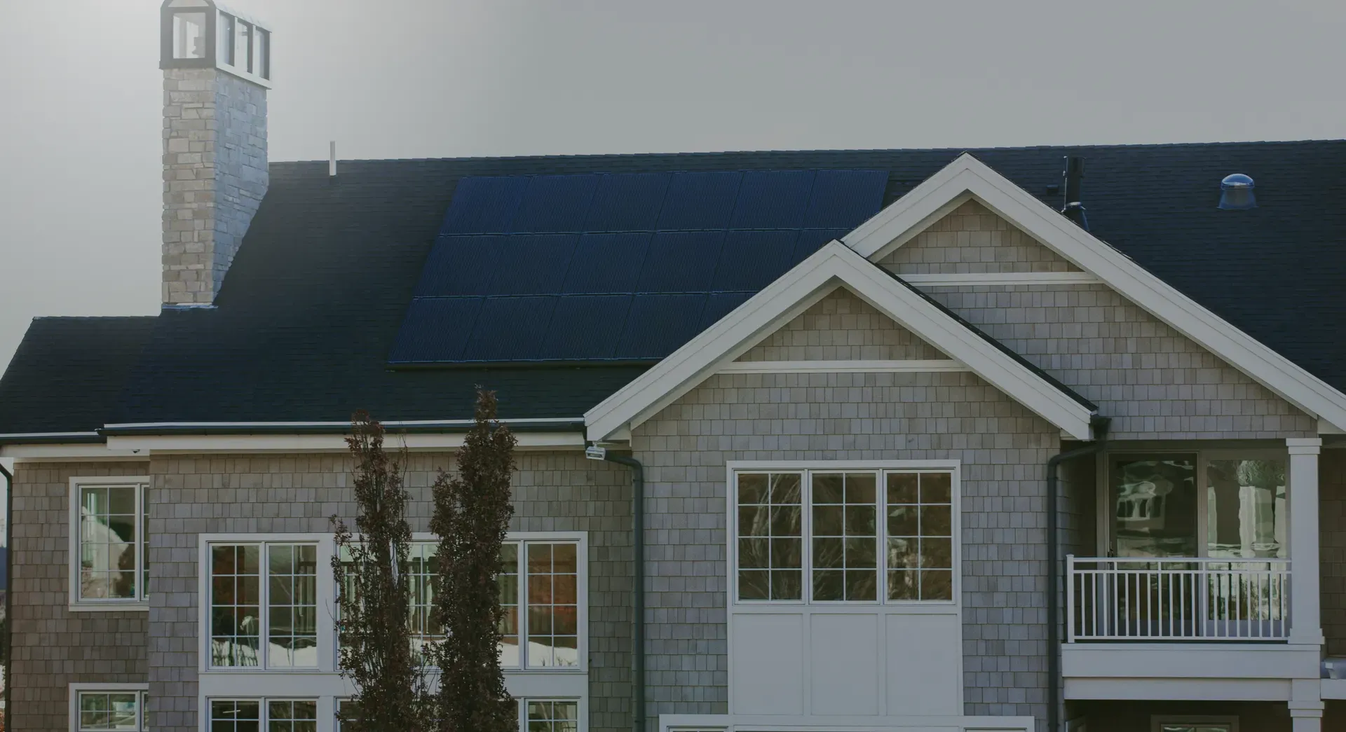 A two-story house with gray siding features solar panels on the dark roof. White-framed windows and a chimney add a modern aesthetic.