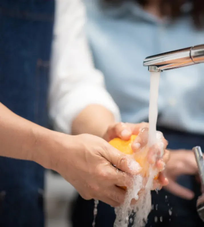 Hands washing an orange under a flowing faucet, conveying cleanliness and freshness. A person in a blue shirt is blurred in the background.