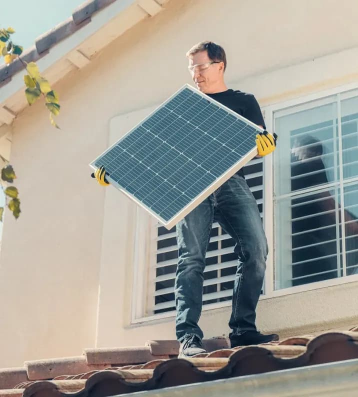 Man on a tiled roof installing a solar panel, wearing safety goggles and gloves. Sunlight emphasizes eco-friendly energy and green living.