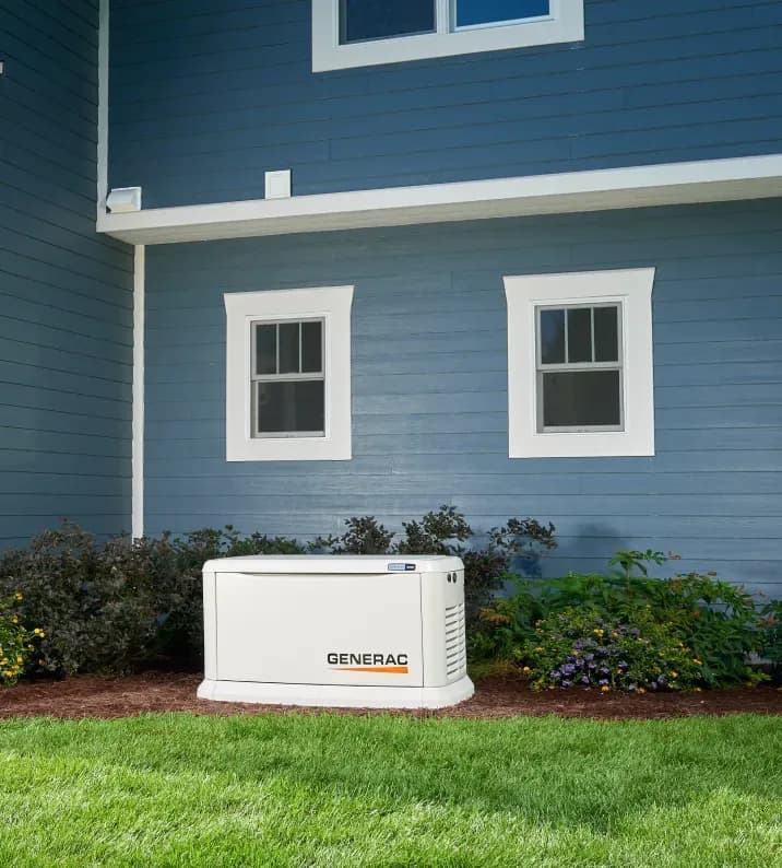A standby generator sits on grass beside a blue house with white trim. Bushes and flowers surround the generator, creating a tidy, well-maintained garden.