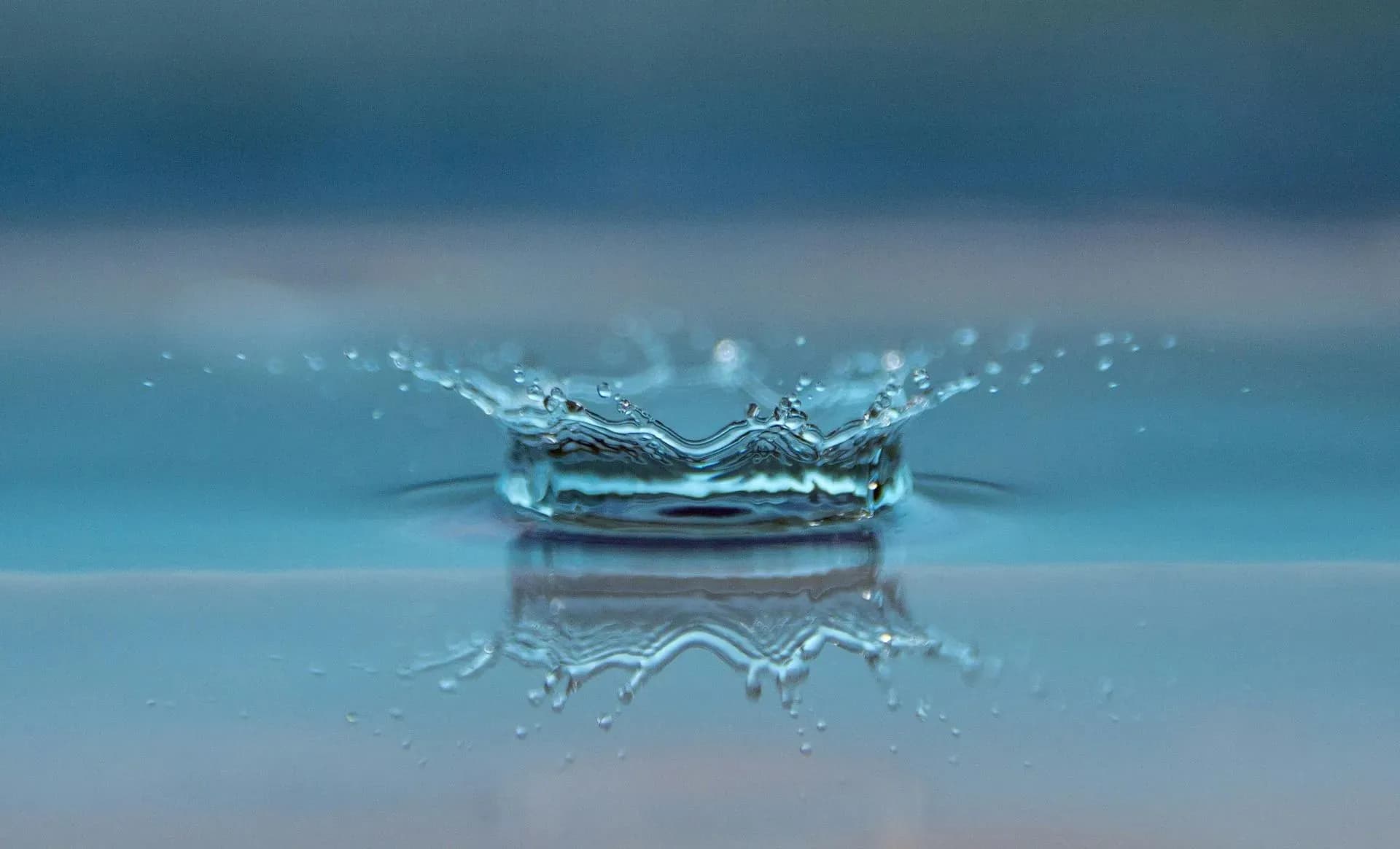 A water droplet creates a splash impact on a calm blue surface, forming a symmetrical crown shape. The image exudes tranquility and precision.