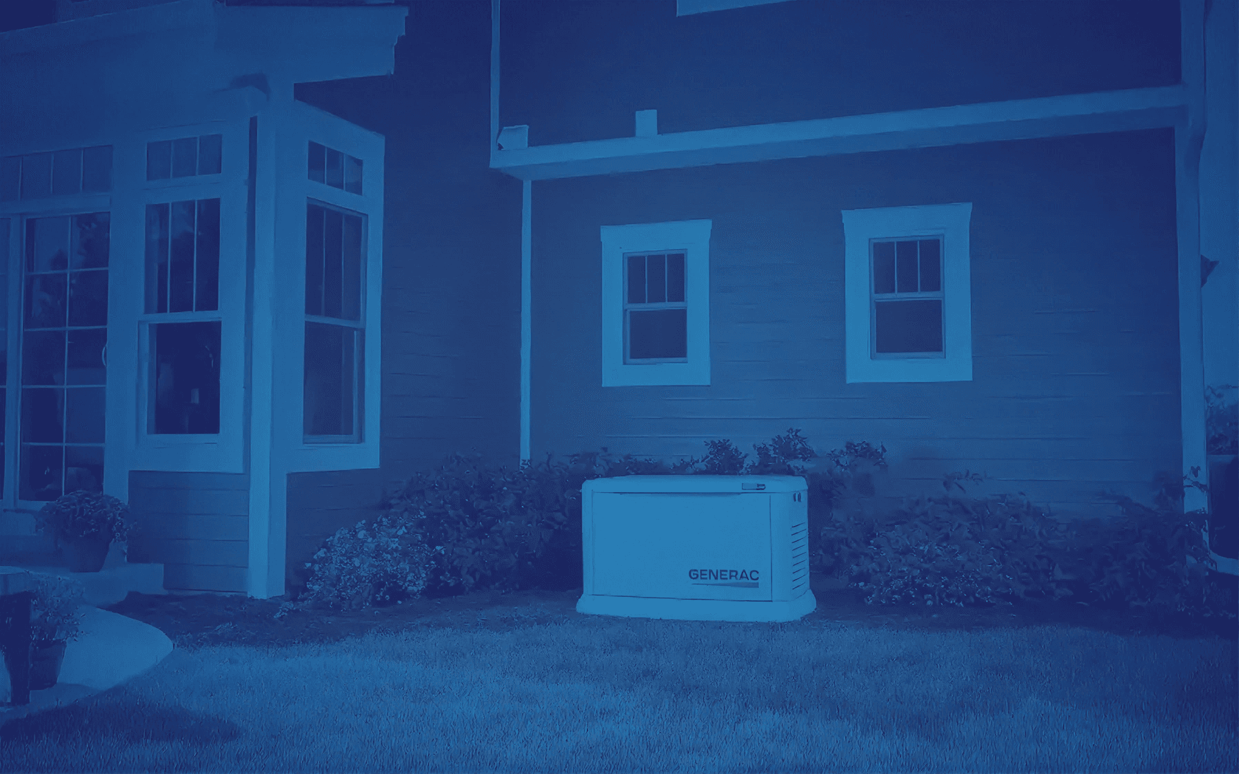 A generator positioned outside a house with siding and two windows. Bushes surround the generator, and large windows are part of an adjacent wall.