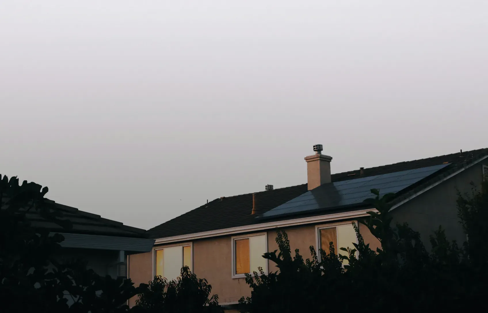 A dimly lit suburban house at dusk, with solar panels on the roof and glowing windows, surrounded by silhouetted trees under a soft, fading sky.