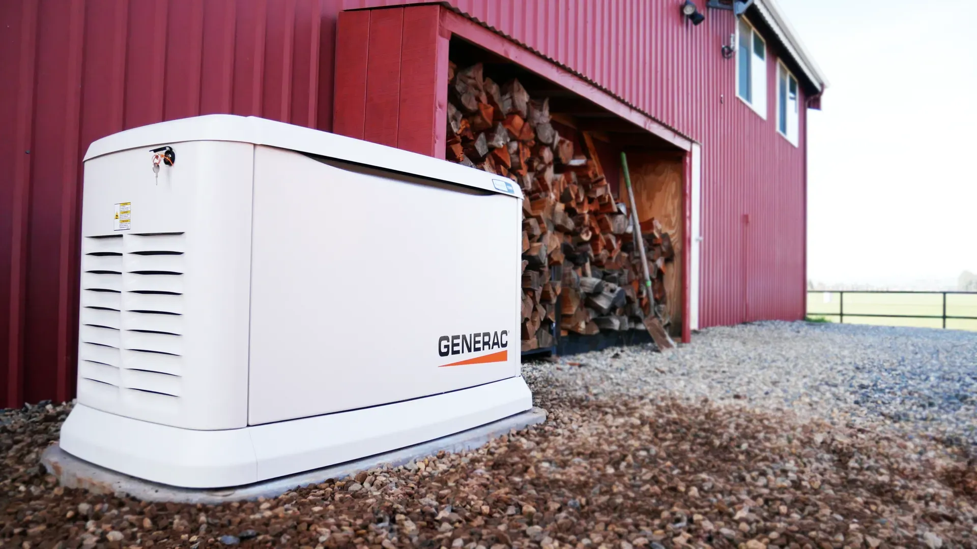 White Generac generator outside a red barn with stacked firewood inside, resting on gravel. The scene conveys preparedness and reliability.