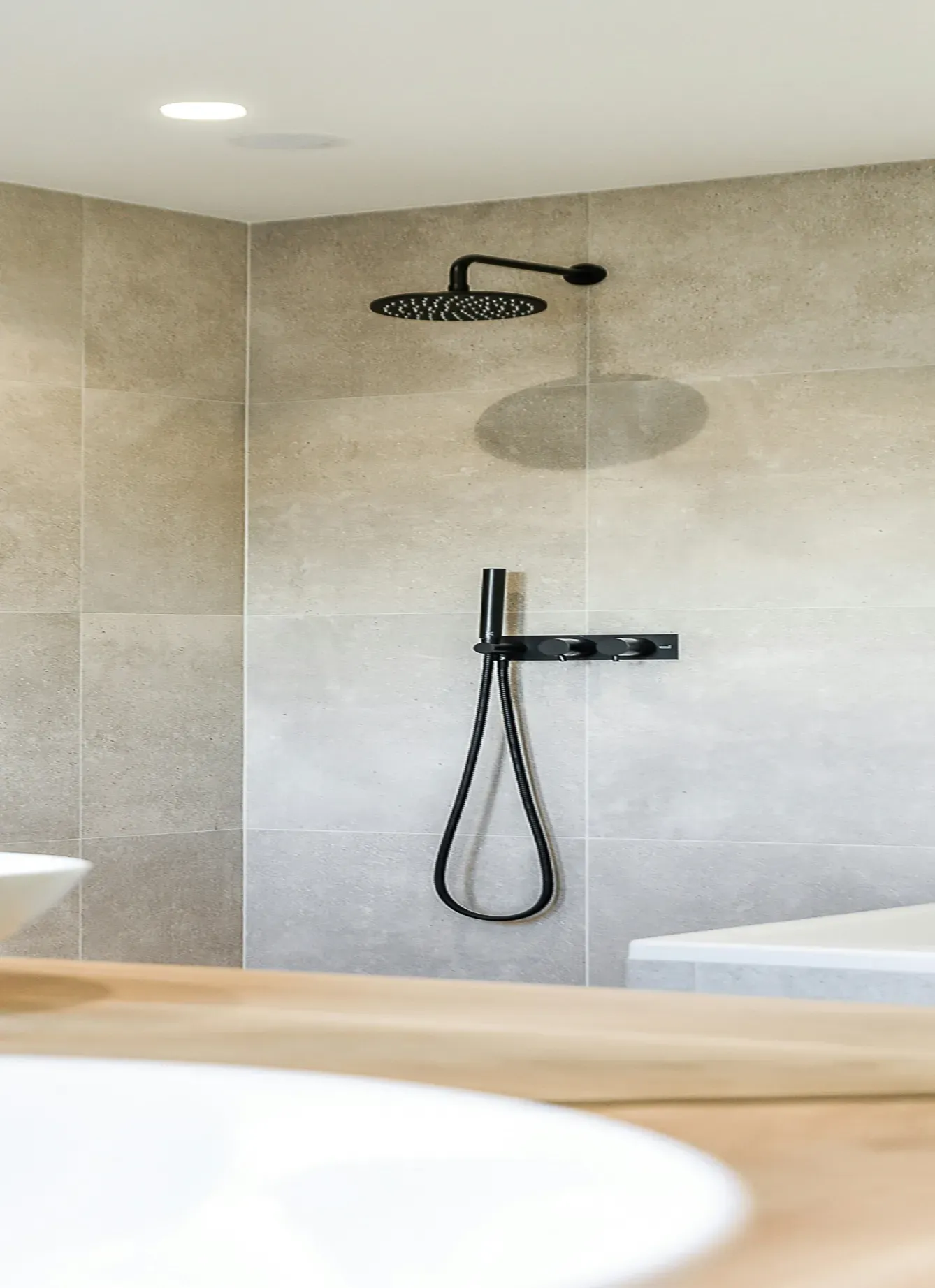 Modern bathroom with beige tiles, featuring a sleek black rainfall showerhead and hose. The tone is calm and minimalist.