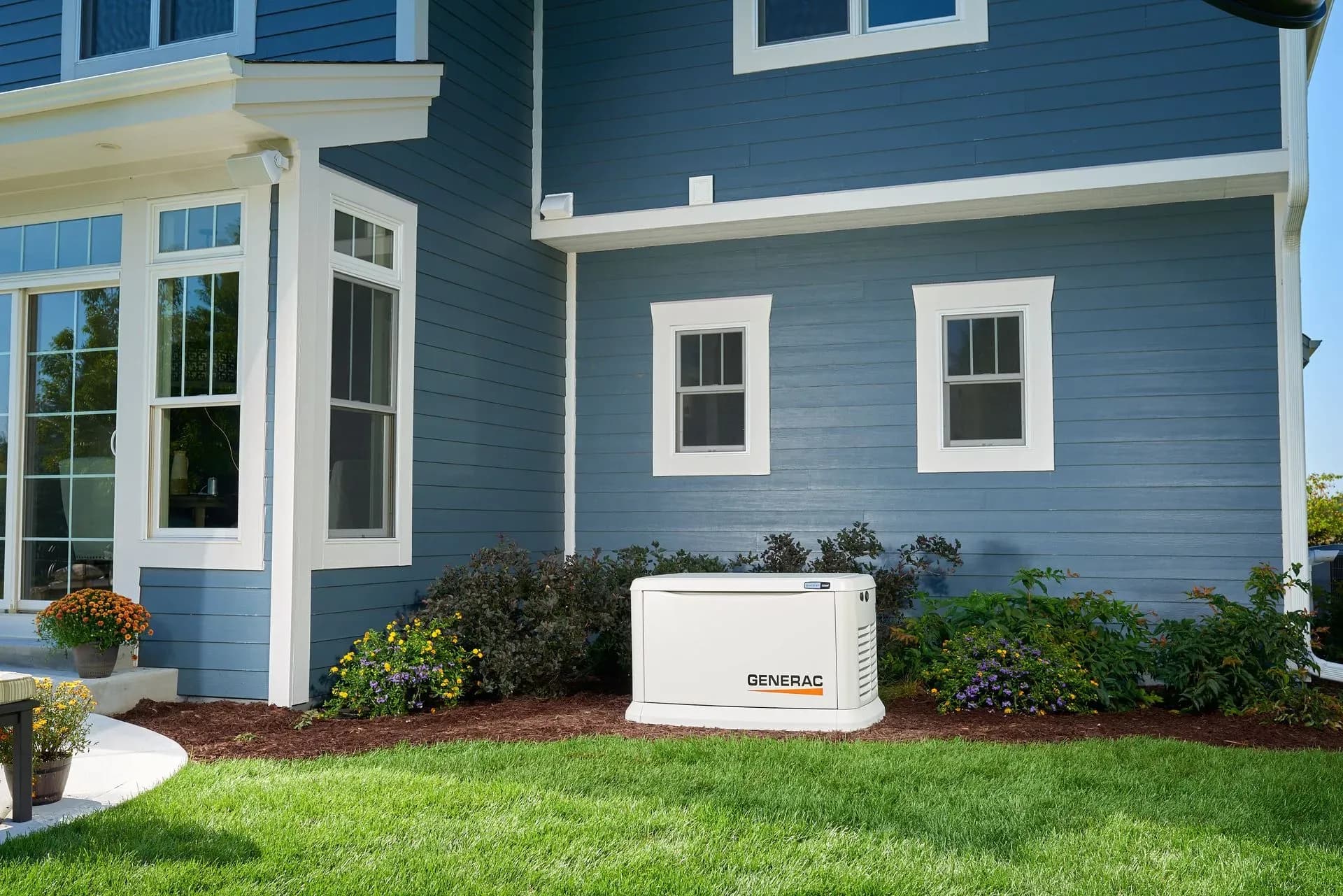 A blue house exterior with white trim is shown in bright daylight. A Generac backup generator is placed on mulch near bushes and greenery, conveying preparedness.