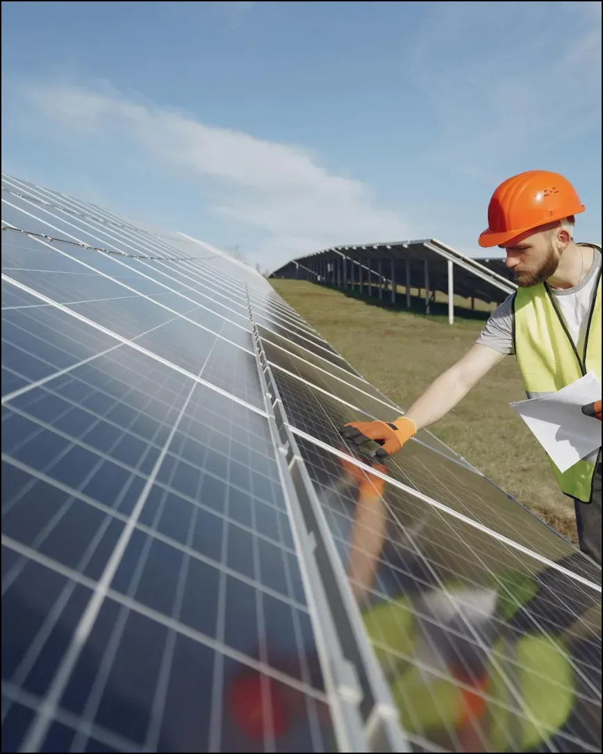 Worker in a hard hat and safety vest inspects solar panels in a field, highlighting sustainable energy. The scene conveys focus and environmental progress.