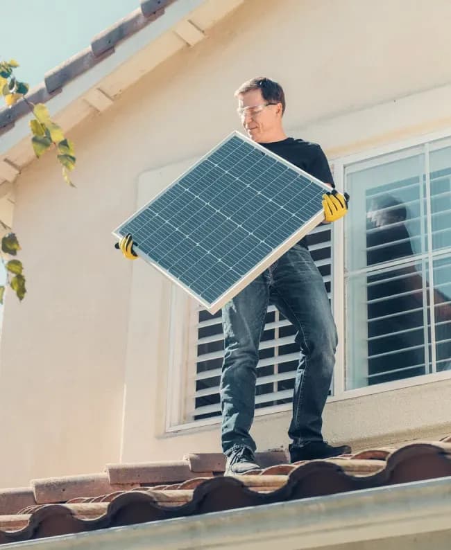 A man in yellow gloves installs a solar panel on a tiled roof in bright sunlight. House window and tree branches visible, conveying eco-friendly action.