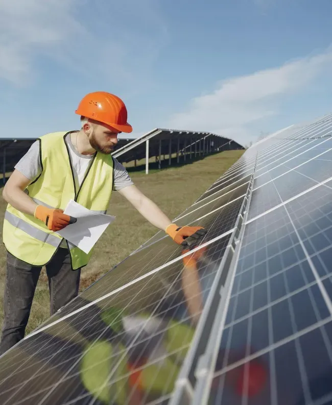 A man wearing an orange hard hat and yellow vest inspects solar panels with a focused expression, under a clear blue sky.