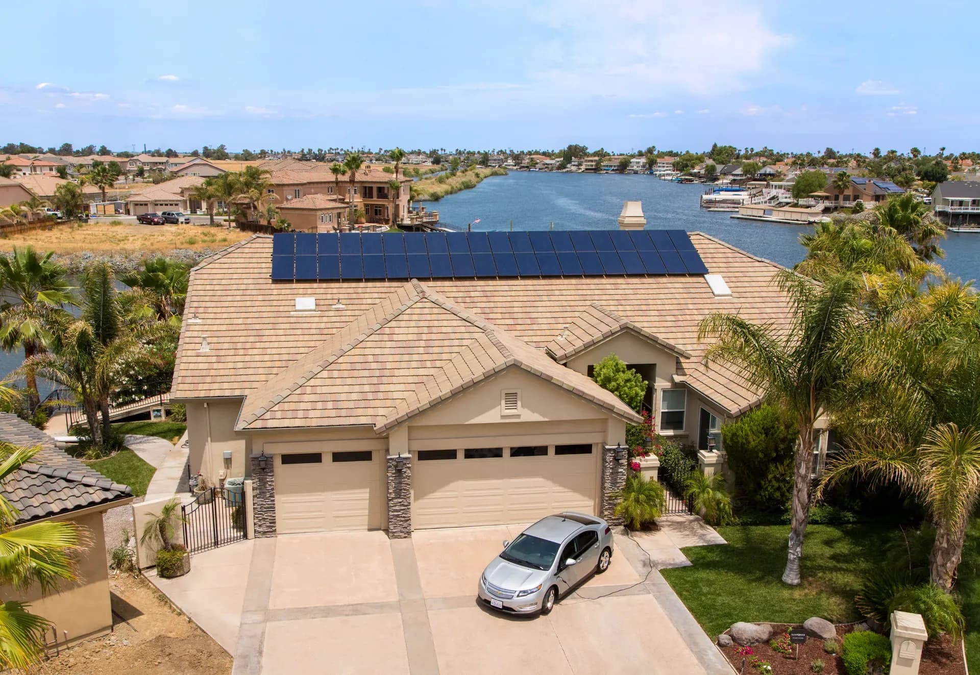 Aerial view of a waterfront house with solar panels on the roof. A silver car is parked in the driveway, surrounded by lush palm trees.