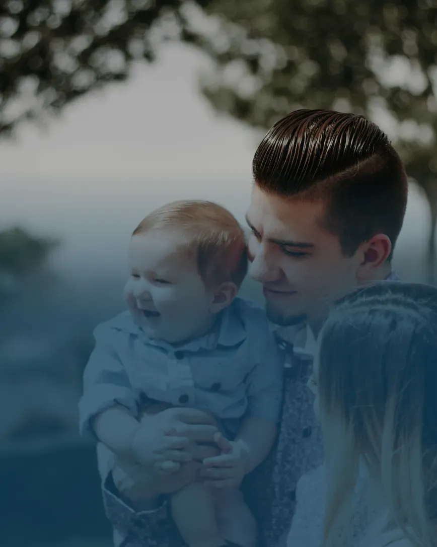 A father, holding a smiling baby in his arms, stands beside a woman outdoors. The scene conveys warmth and joy, with a blurred forest background.