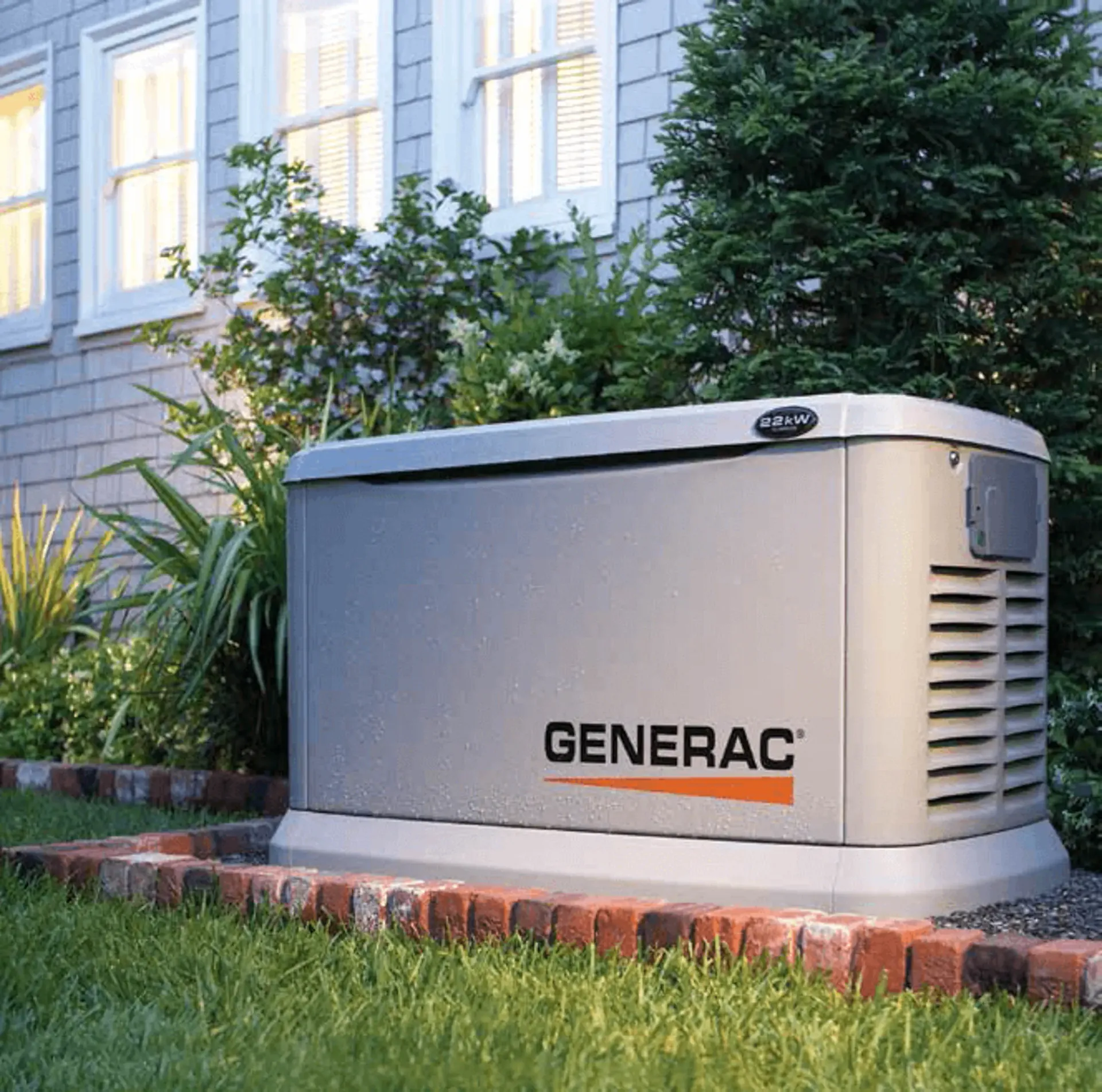 A Generac generator in a garden next to a house with lit windows. It's surrounded by greenery and sits on a brick-bordered platform. The scene is peaceful.