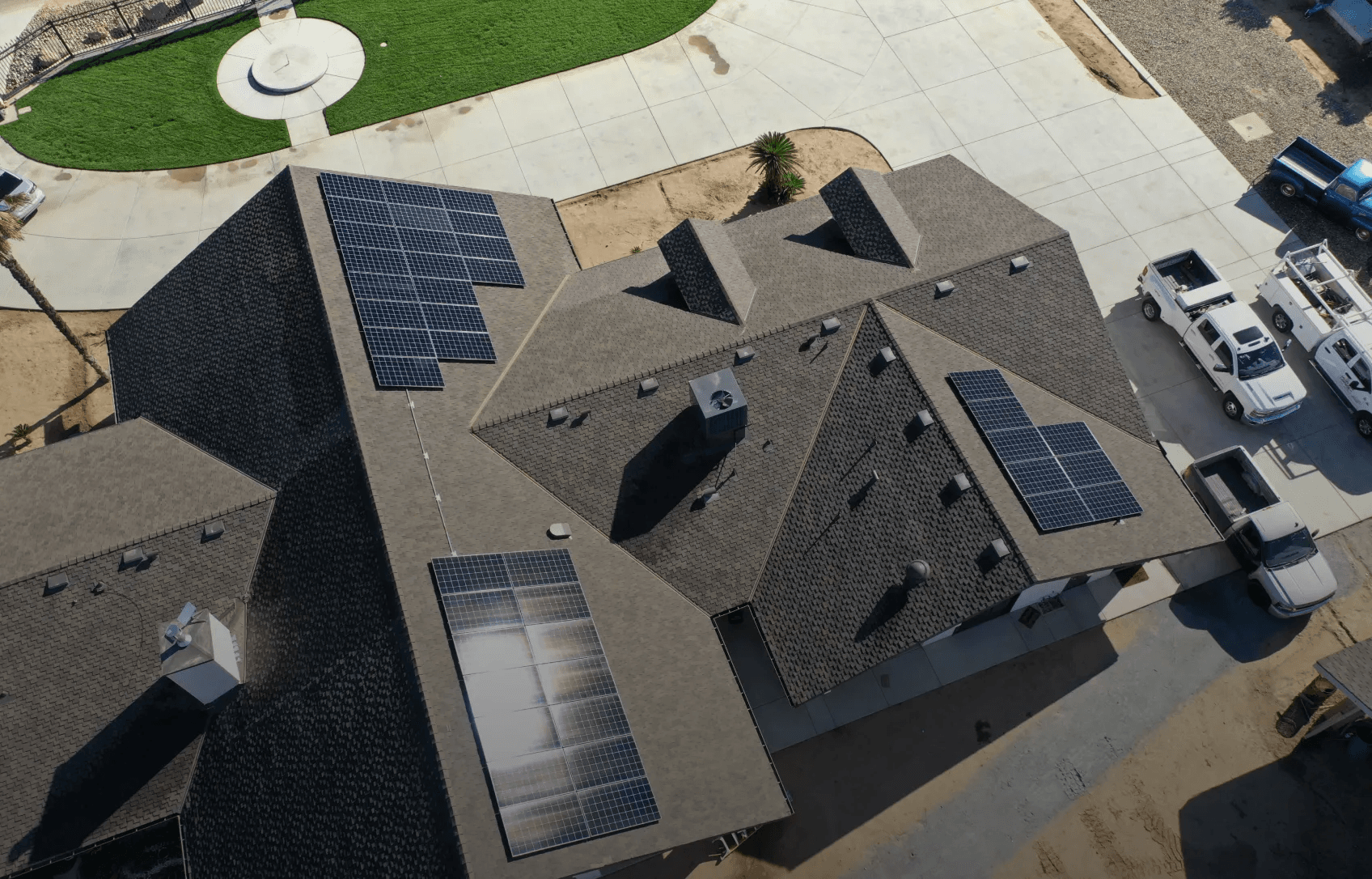 Arial view of a house with multiple solar panels on its roof. Nearby, several parked white trucks and a small grassy area are visible, creating a modern and eco-friendly atmosphere.