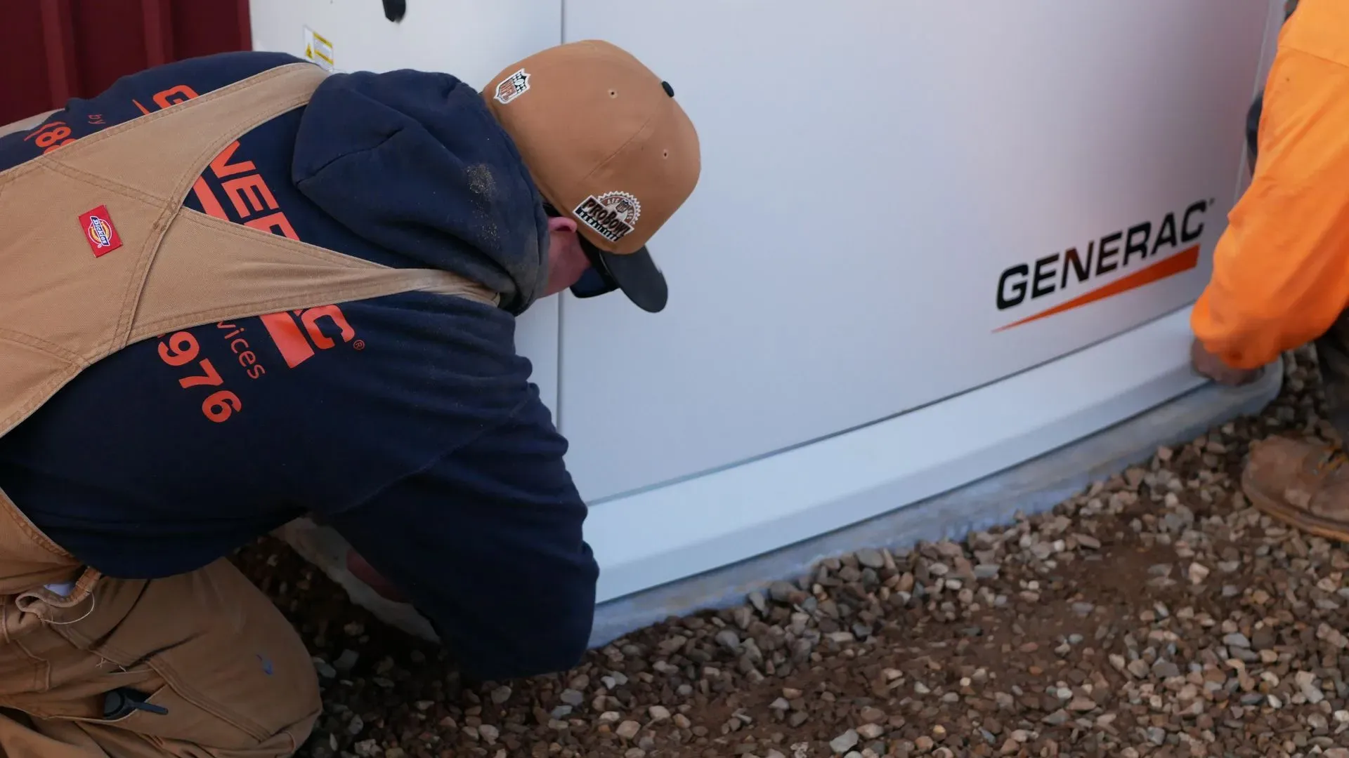 Two workers in outdoor gear install a Generac generator on gravel. They are focused, kneeling, and adjusting its base, indicating attention to detail.