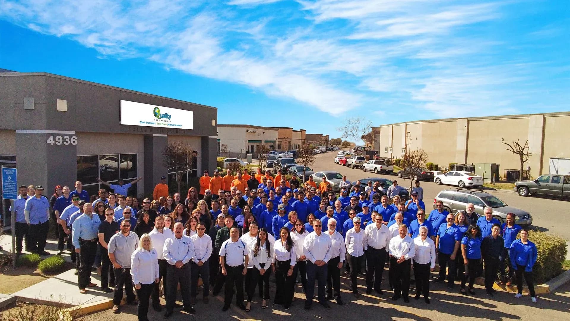 A large group of people in colorful shirts gather smiling outside an office building under a blue sky, conveying a sense of community and teamwork.