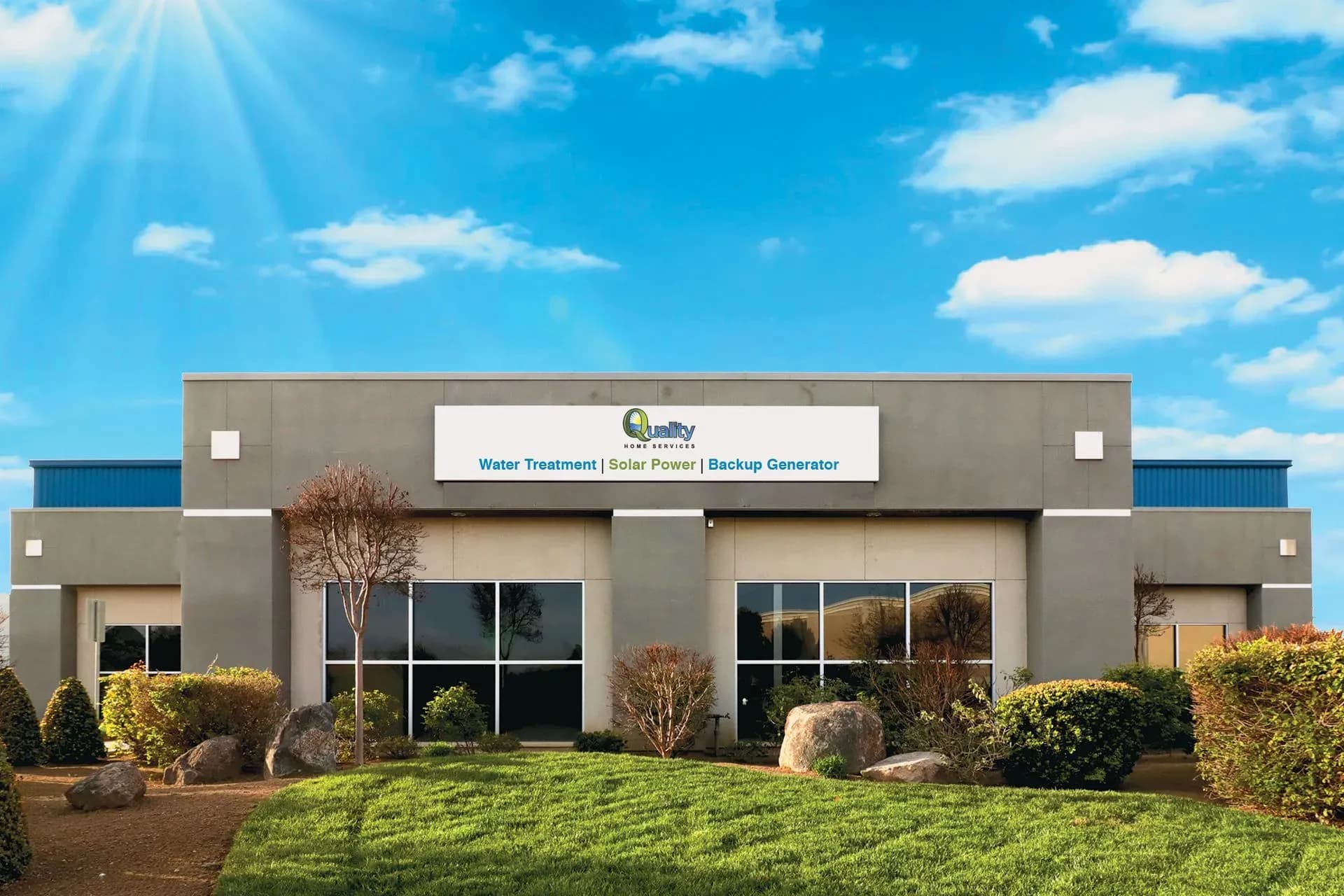 Modern building under a bright blue sky with scattered clouds. Sign on facade reads "Water Treatment | Solar Power | Backup Generator". Front landscape includes neatly trimmed bushes and rocks.