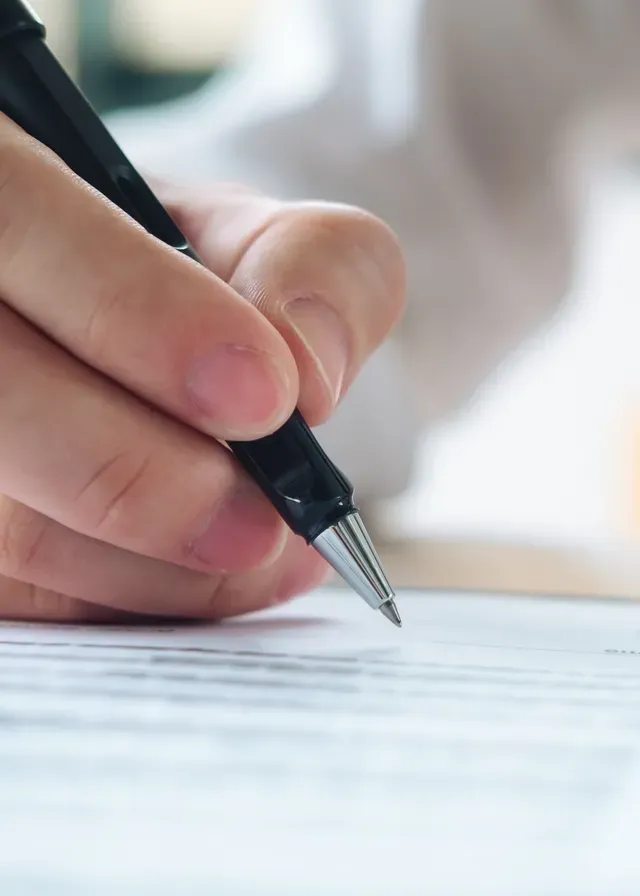 Close-up of a hand holding a black pen, writing on a document. The focus is on the pen tip, conveying concentration and precision.