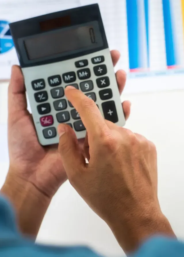 Close-up of hands using a calculator, pressing buttons, with a blurred background of colorful graphs. Focus on calculation and analysis.