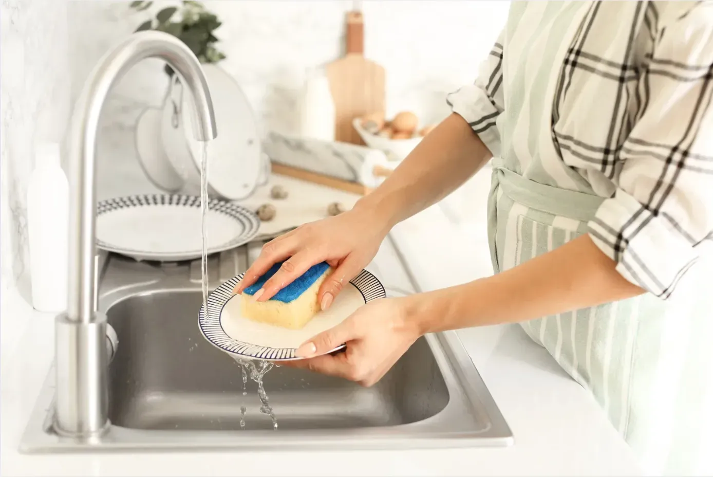 Hands washing a plate with a blue sponge under running water in a modern kitchen. Sunlit sink area with cutting board and eggs in background.
