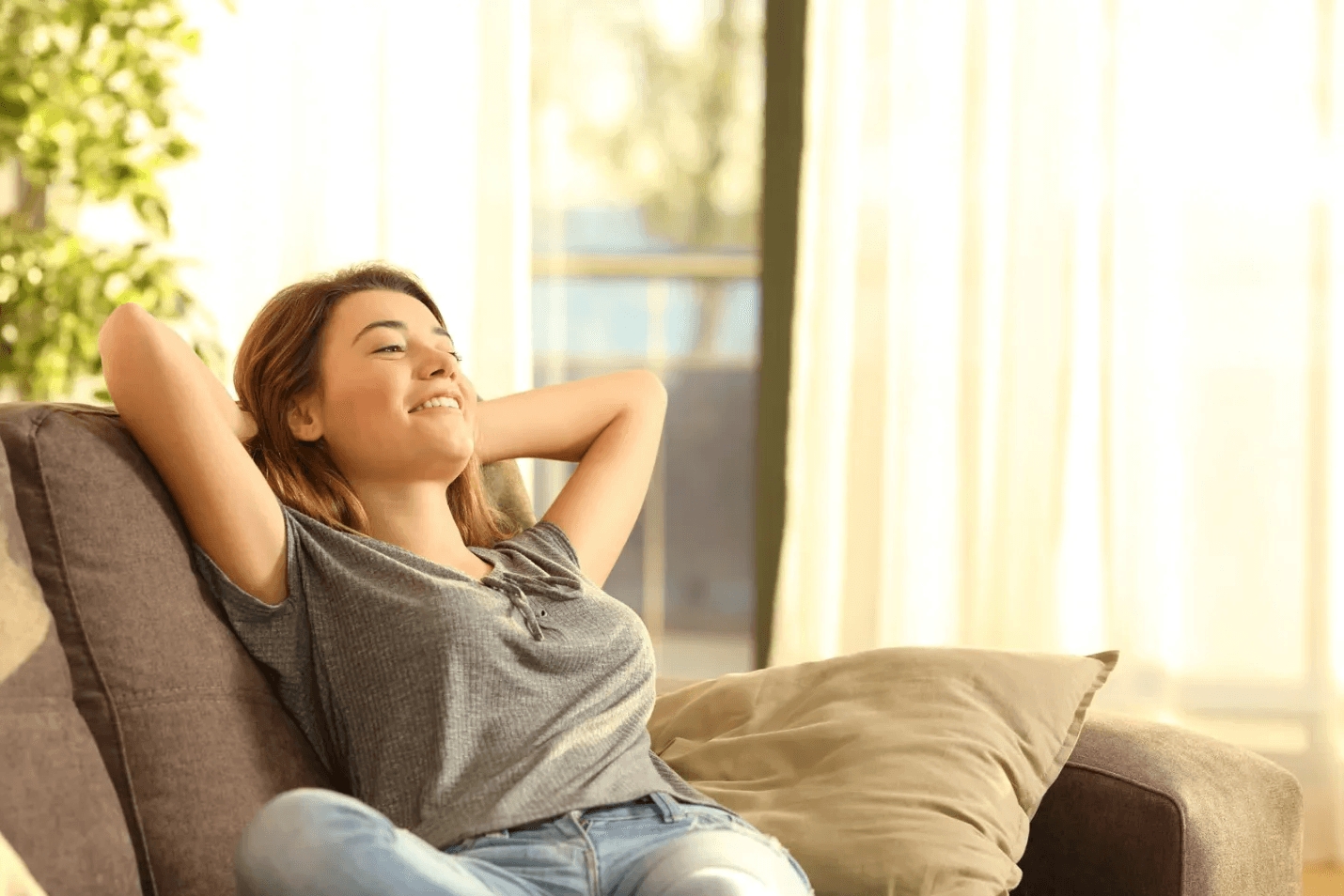 Woman relaxing on a sofa, arms behind her head, smiling peacefully. Sunlight streams through sheer curtains, creating a warm, tranquil atmosphere.