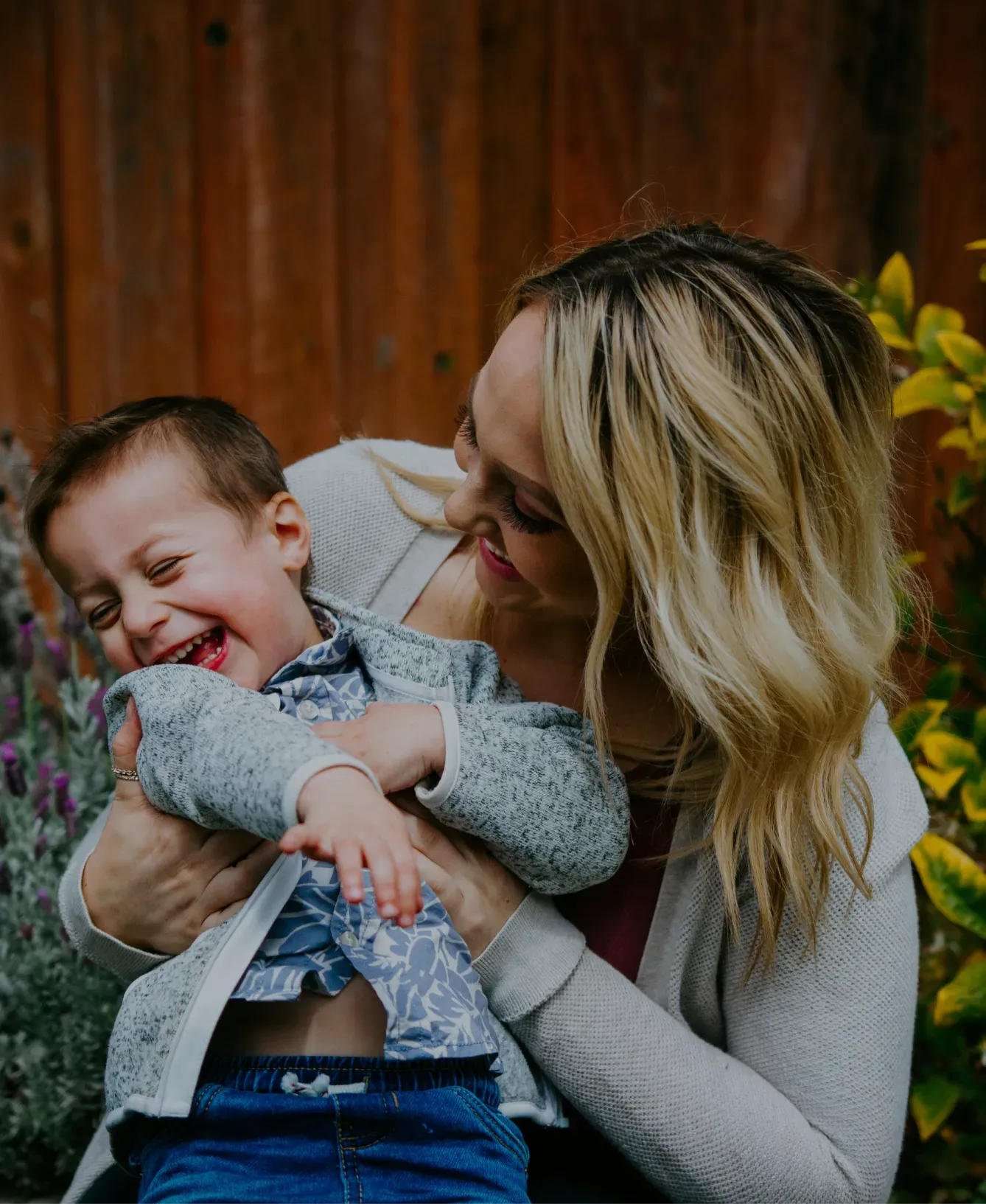 A woman with blond hair joyfully hugs a laughing toddler in a blue shirt and gray sweater, set against a wooden fence and greenery.