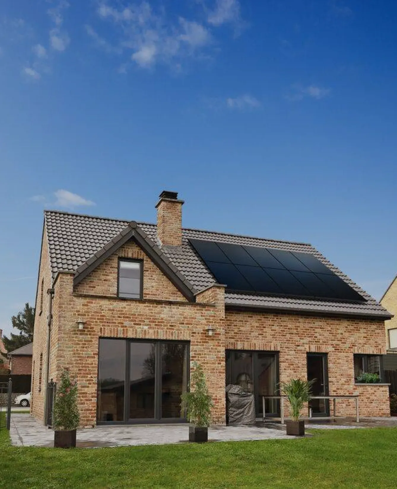 Red brick house with solar panels on the roof, large glass doors, and a neatly landscaped garden. The sky is clear and blue.