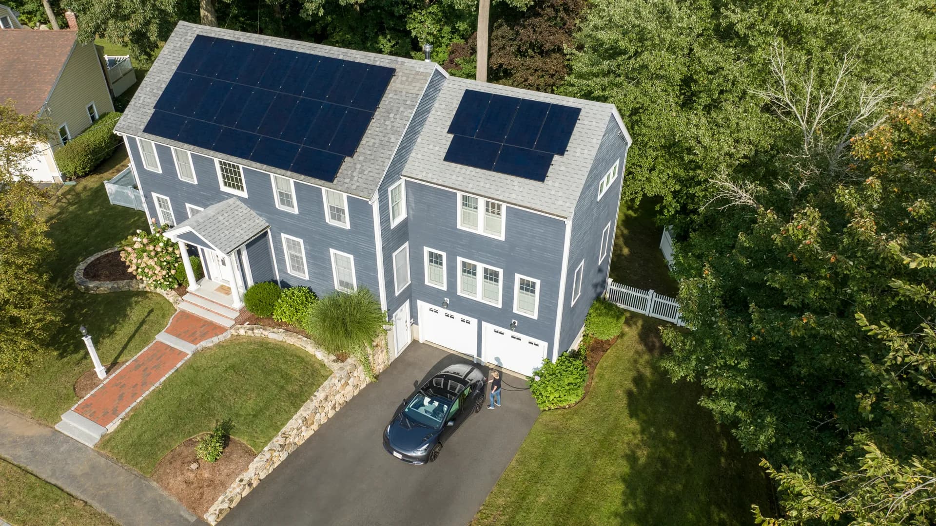 Aerial view of a large blue house with solar panels on the roof. A couple stands beside a car in the driveway, surrounded by green lawns and trees.
