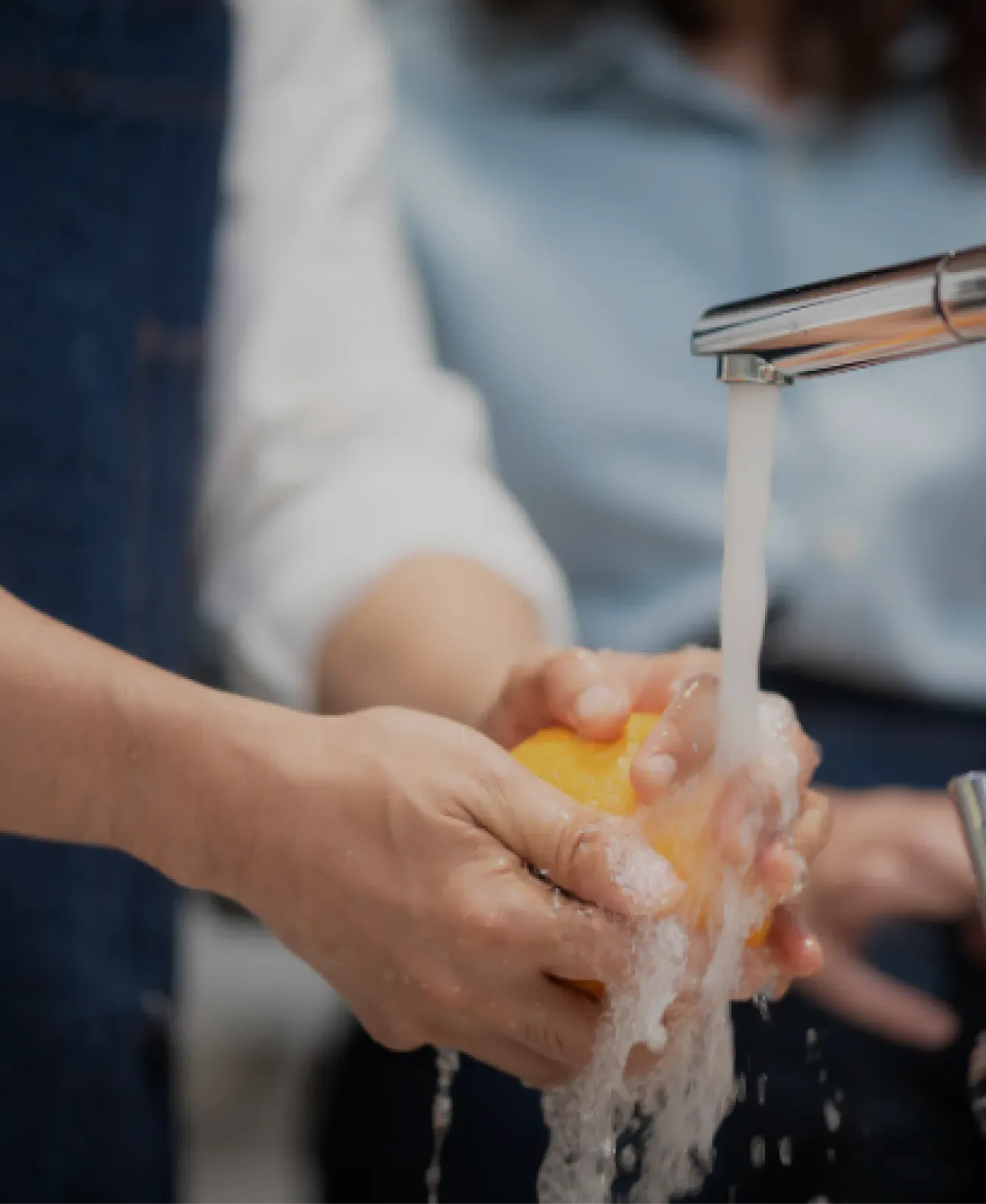 Two people wash a yellow bell pepper under a running kitchen faucet. One person wears an apron, suggesting a cooking activity, with a collaborative vibe.