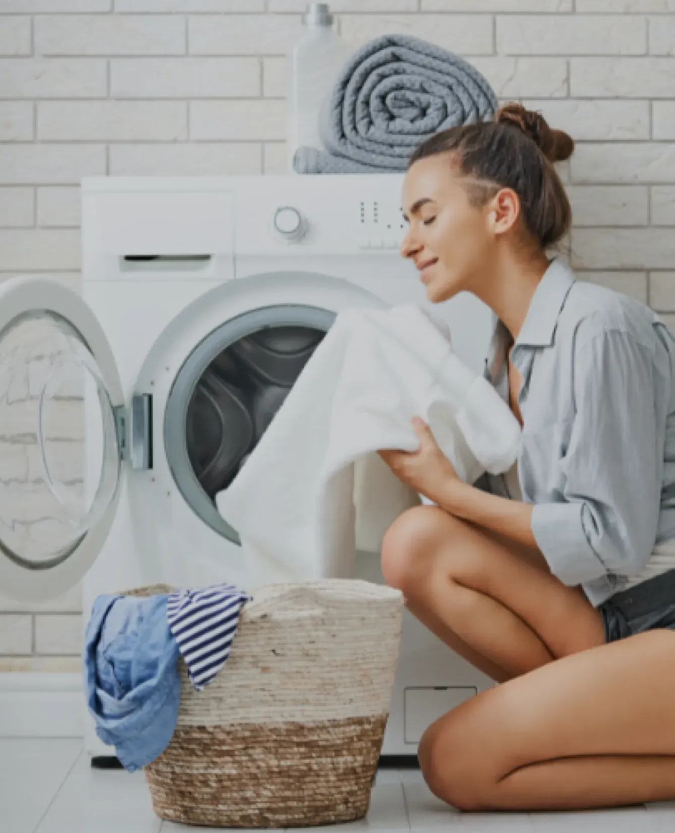 Woman kneeling by an open washing machine, smiling, holding a white towel. Basket with clothes beside her; neatly folded towels atop the machine. Cozy and clean atmosphere.
