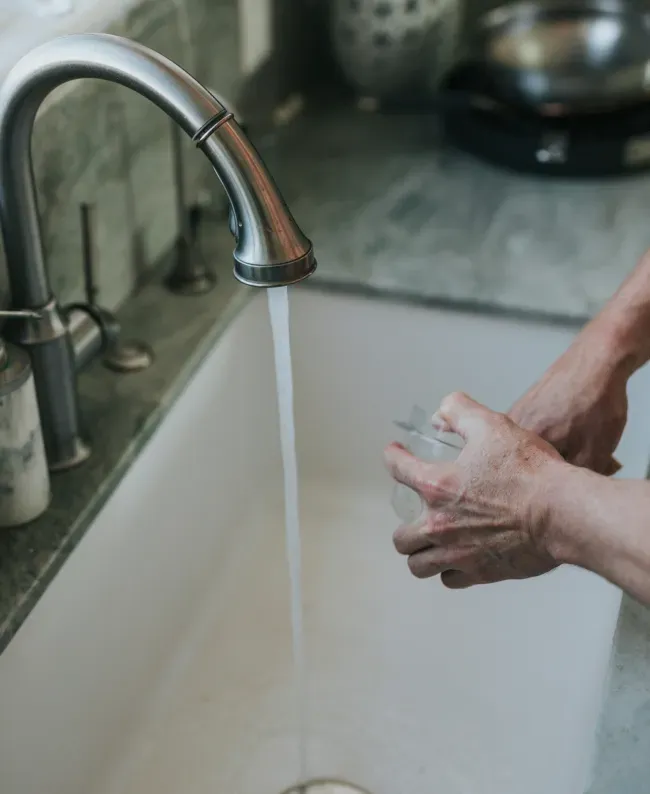 Hands washing a glass under a steady stream of water from a kitchen faucet. The setting is calm, with a gray countertop and a white sink.