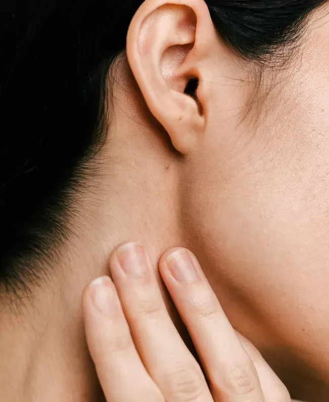 Close-up of a person touching their neck below the ear with fingers. The skin appears smooth, and the dark hair contrasts against the light skin tone.