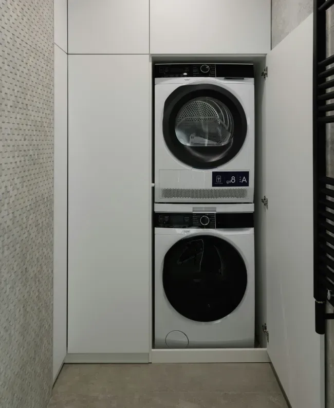 Stacked washer and dryer inside a sleek, white cabinet with open doors, set against a textured wall. The space is modern and minimalist.