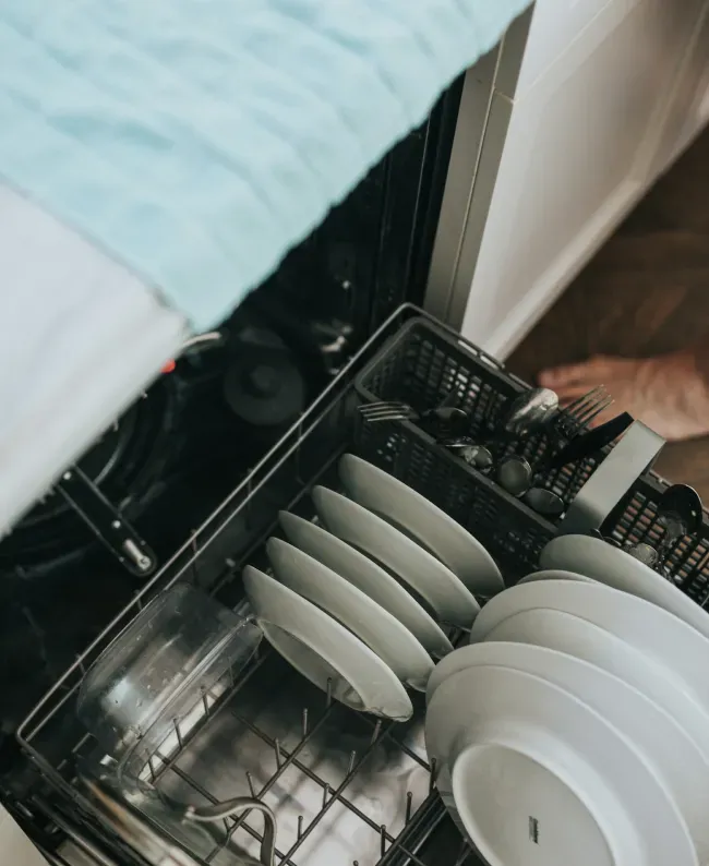 Open dishwasher with neatly stacked clean white plates and cutlery. A person’s hand is visible near the dishwasher, suggesting tidiness and routine.