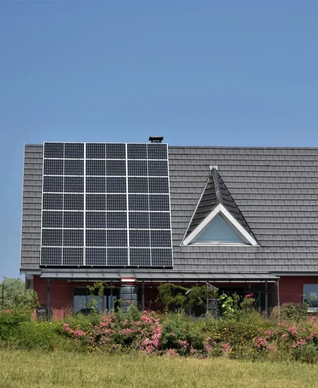House with solar panels on a tiled roof, set against a clear blue sky. A garden with pink and green foliage adds vibrant contrast at the front.